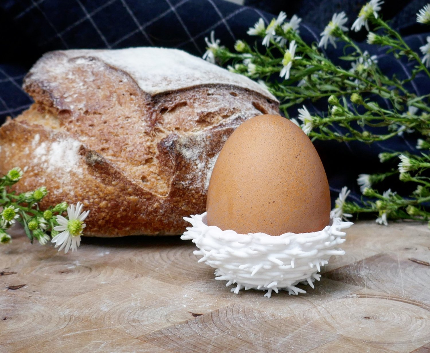 3D printed white egg nest with brown egg inside on a wooden table with flowers and loaf of bred